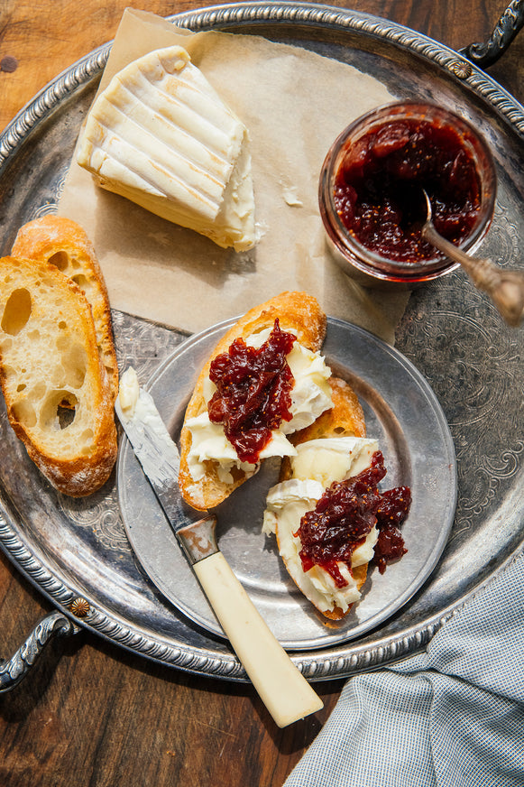 An overhead styled food scene on a large silver tray, showing a slice of white cheese, a jar American Spoon Holiday Jam with a spoon, two pieces of baguette, and two open-faced bread slices topped with cream cheese and the jam, with a knife resting on the tray.