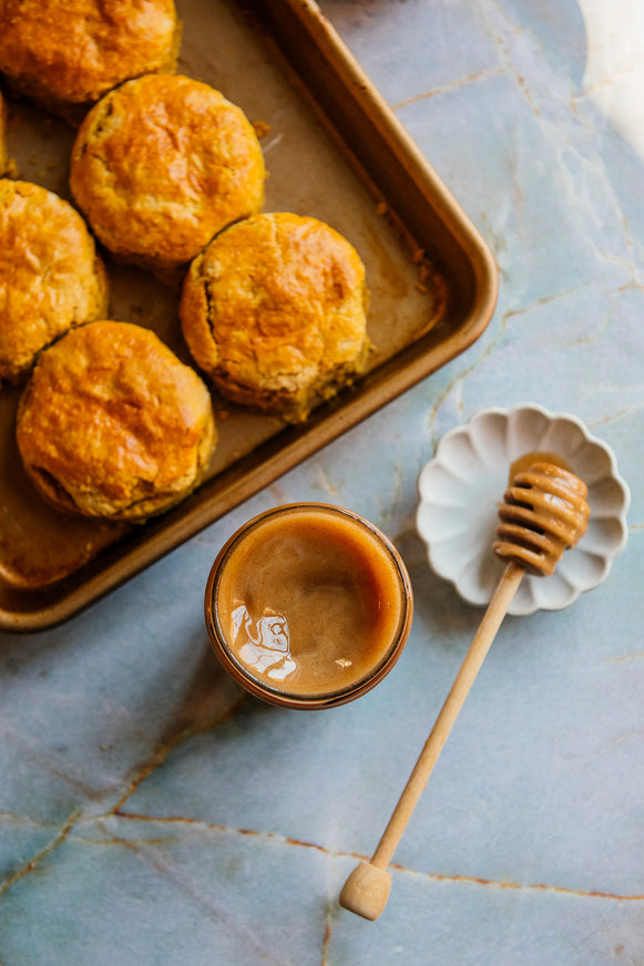 Biscuits on a sheet pan with a jar of Cinnamon Honey and a honey dipper on a marble surface.
