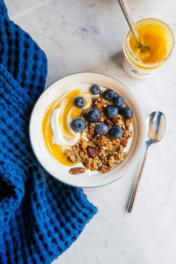 A bowl of yogurt topped with lemon curd, blueberries and maple granola next to an open jar of Lemon Curd and a blue tea towel