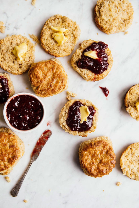 Overhead of several golden-brown baked biscuits on a white marble surface, with some sliced open and topped with butter and strawberry preserves A small white bowl of jam and a knife are also visible.