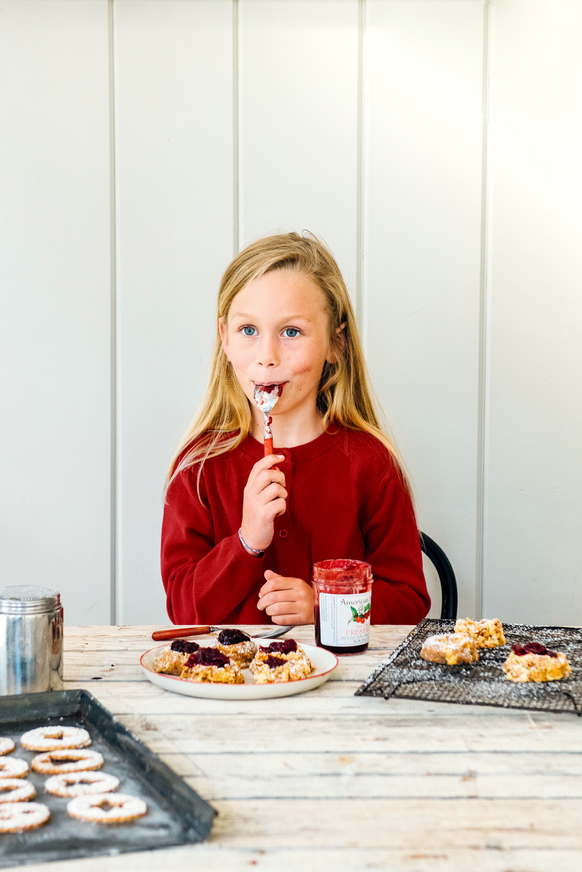 A young girl eating a spoon full of preserves while making thumbprint cookies