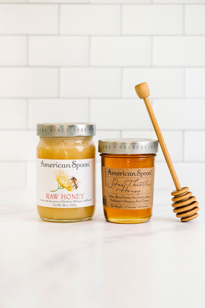 A jar of Raw Honey and Star Thistly Honey, with a honey dipper leaning against one jar