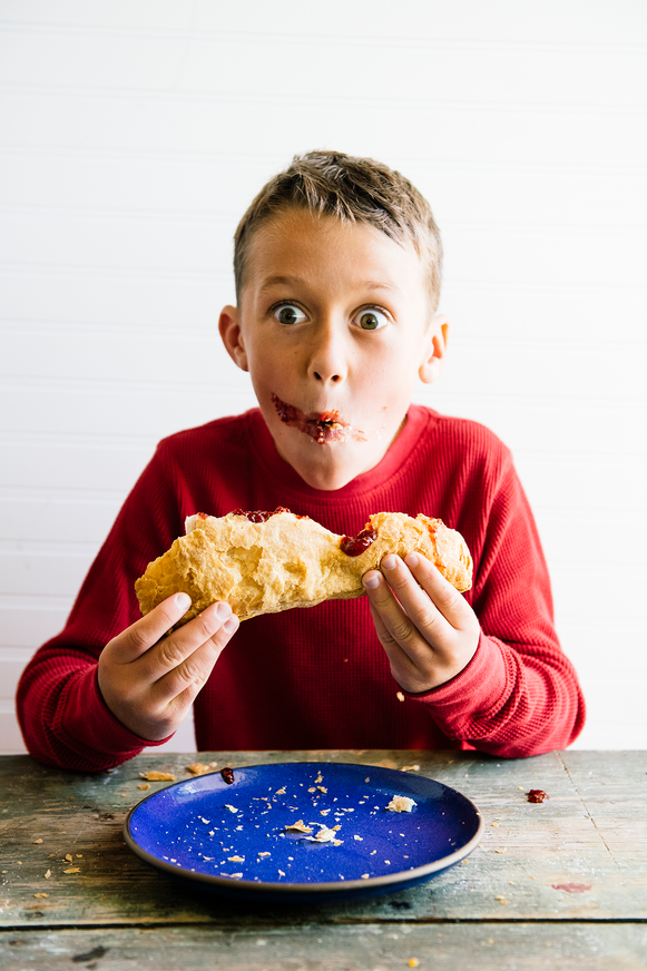 A young boy pulling apart a jam filled croissant with jam across his cheek