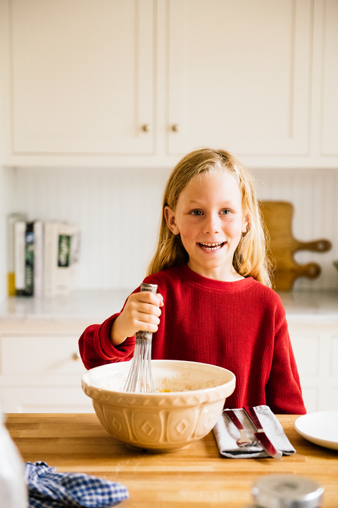 Load image into Gallery viewer, A young girl at a butcher block kitchen counter, mixing pancake mix in a large bowl with a whisk
