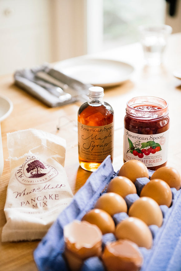 A kitchen counter filled with Wheat & Malt Pancake Mix, Maple Syrup, a carton of eggs and Fruit Perfect Sour Cherries